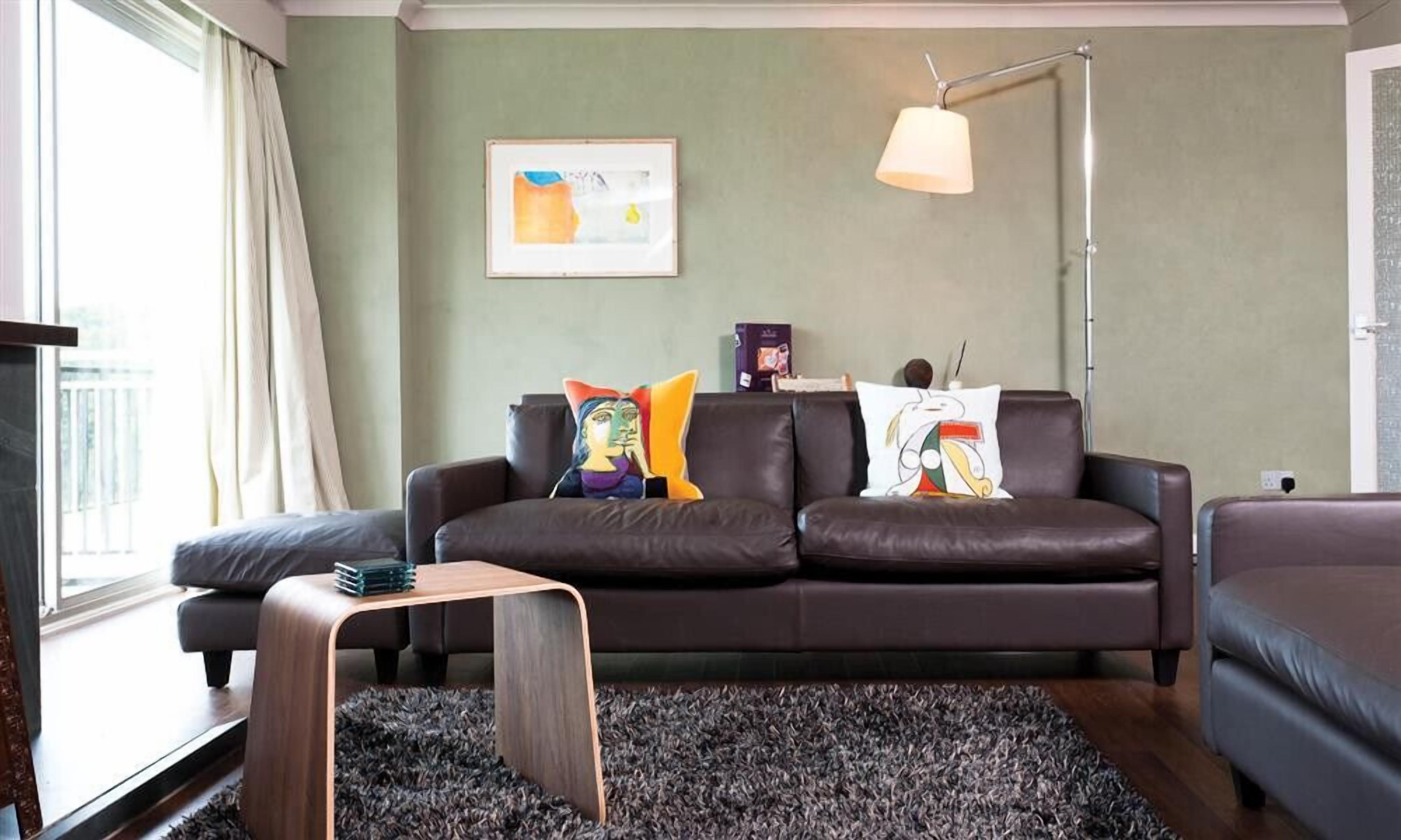 Un salon moderne avec un canapé en cuir marron foncé, une petite table d’appoint en bois, un tapis noir à longues mèches, un lampadaire et un tableau abstrait encadré accroché au mur.