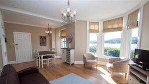 A living room with a brown sofa, a dining table with chairs, a TV, and a view of the outdoors through large windows. - Waterside (Ambleside)