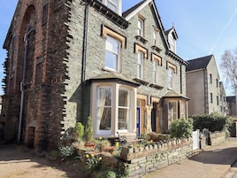 A two-story stone house with a bay window, a front door, and a small garden with flowers.
