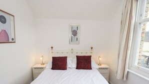 A neatly made bed with white linens and two red pillows, flanked by bedside tables with lamps, in a room with a simple white wall and a framed picture above the bed. - Number Twenty (Keswick)