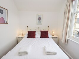 A neatly made bed with white linens and two red pillows, flanked by bedside tables with lamps, in a room with a simple white wall and a framed picture above the bed.