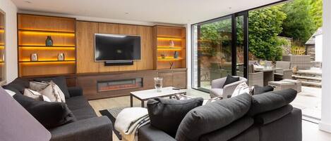 A modern living room with a flat-screen TV, built-in shelves, and a large window overlooking a garden.