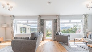 Een ruime woonkamer met een moderne grijze bank, een kleinere fauteuil en een houten salontafel. Grote ramen bieden uitzicht op het omliggende landschap.