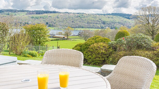 Een ronde tafel met rieten stoelen en twee glaasjes sinaasappelsap, met uitzicht op een mooi landschap met heuvels en een meer.