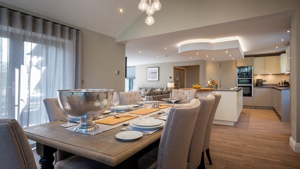 A modern dining area with a large table set for six, a kitchen in the background, and a view of the outdoors through large windows. - Landower House (Ambleside)