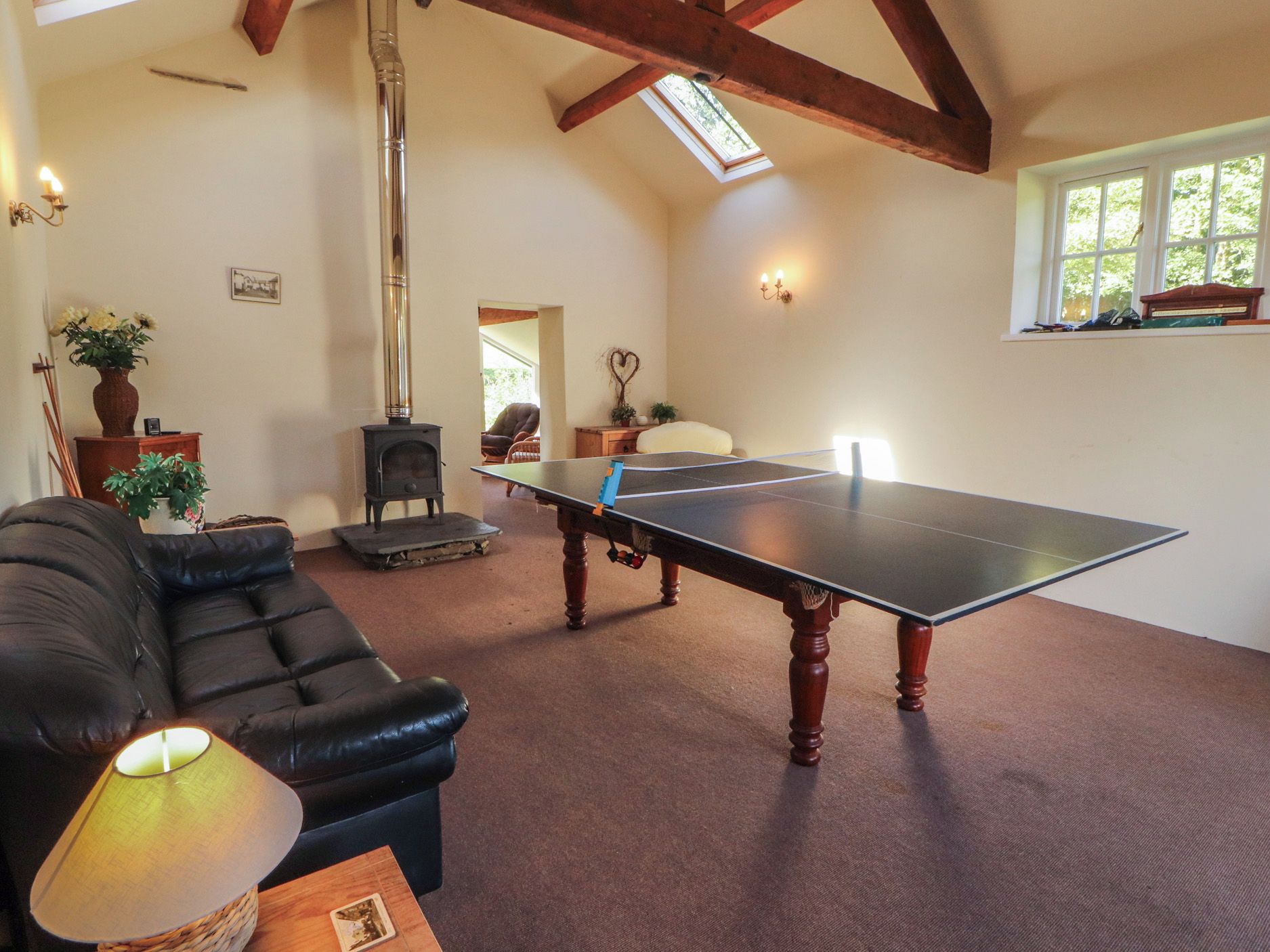 A room with a ping pong table, a sofa, a lamp, a vase with flowers, a wooden table, and a fireplace.