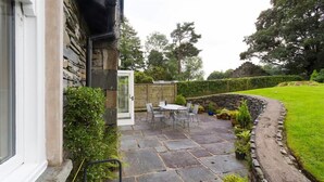 Un patio de piedra con una mesa y sillas, rodeado por un muro de piedra y una zona ajardinada.