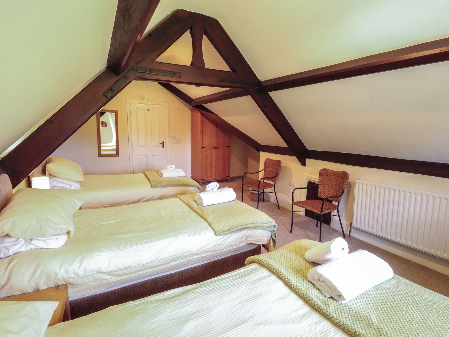 A room with a sloped ceiling, wooden beams, a bed with linens, a chair, and a radiator.
