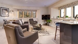 A modern living room with leather sofas, a coffee table, and a TV. There is a dining area with a bar and a large window. - Hygge at No.8 (Windermere)