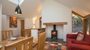 A cozy living room with a wooden dining table, chairs, a red sofa, a black wood-burning stove, and a large window. - Spring Cottage (Ambleside)