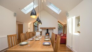 A dining area with a wooden table set for six, flanked by wooden chairs. A fireplace is visible in the background, and the room has a sloped ceiling with skylights. - Spring Cottage (Ambleside)