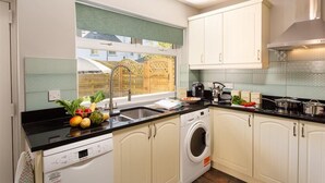 A modern kitchen with a dishwasher, washing machine, and a window with a view of a wooden fence. - Little Leveret Cottage (Keswick)