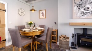 A dining area with a round wooden table, grey upholstered chairs, a wall clock, a framed picture, and a fireplace with a black stove. - Little Leveret Cottage (Keswick)