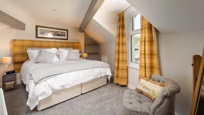 A bedroom with a bed, a nightstand, a chair, a window with curtains, and a painting on the wall. - Little Leveret Cottage (Keswick)
