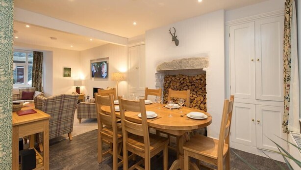 Un coin repas avec une table ronde en bois, dressée pour six personnes, entourée de chaises en bois. On aperçoit au fond une cheminée avec un manteau en pierre.