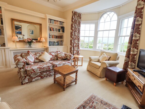 A cozy living room with a floral sofa, a wooden coffee table, a beige armchair, and a bookshelf filled with books. - Grisedale View (Keswick)