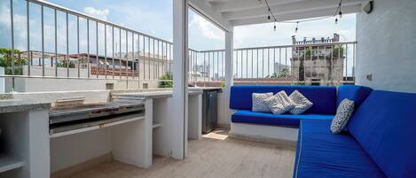 A rooftop terrace with a blue sofa, a grill, and a view of buildings in the background.