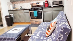 Une cuisine compacte avec des meubles gris, un four à micro-ondes et une cuisinière. On y trouve une petite table à manger recouverte d’une nappe à carreaux bleus et blancs.