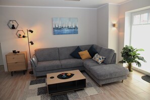Un salon moderne avec un canapé d’angle gris, une table basse en bois, une petite table d’appoint et un tableau représentant des voiliers accroché au mur.