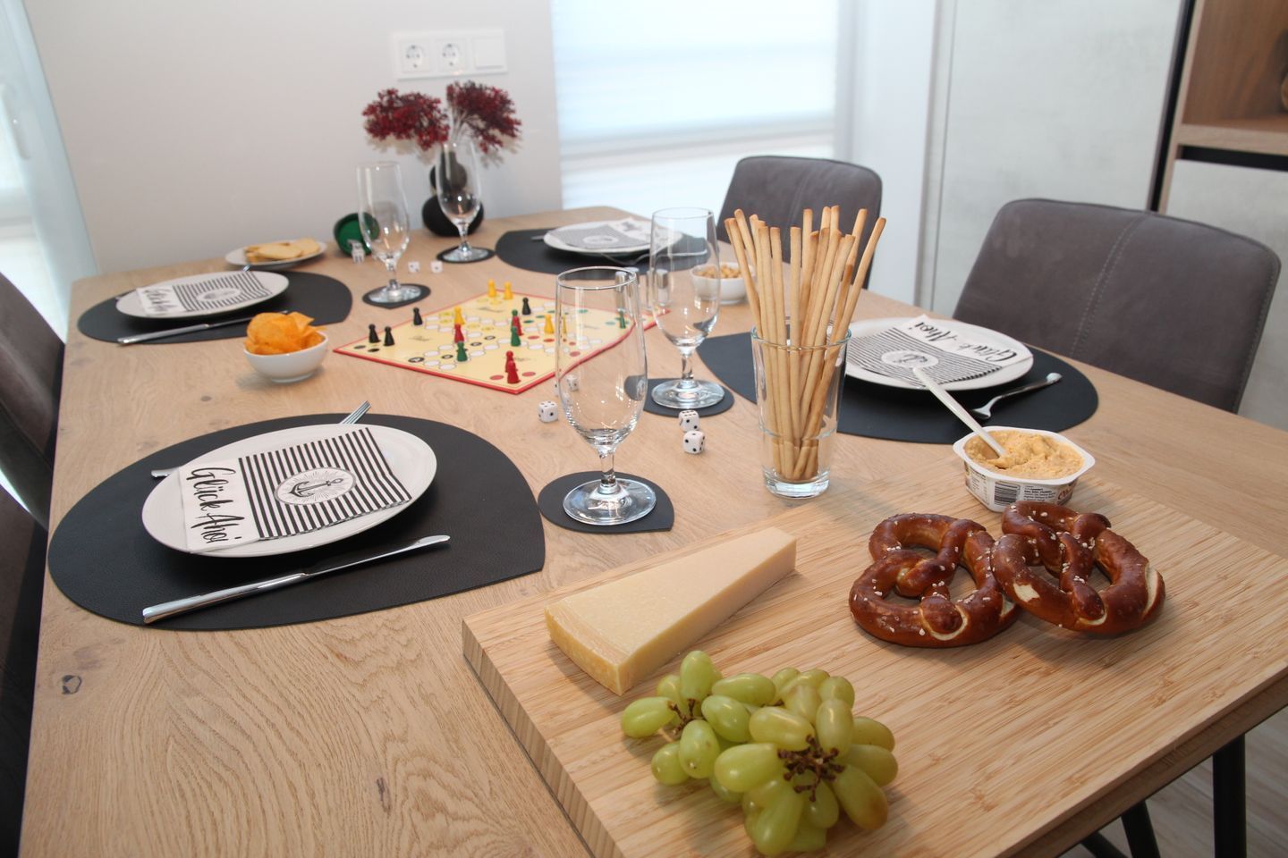 Una mesa de comedor dispuesta con platos, cubiertos, vasos, un tablero de ajedrez, un jarrón con flores, un bol de aperitivos, vasos y botellas de agua, y vasos y botellas de vino.