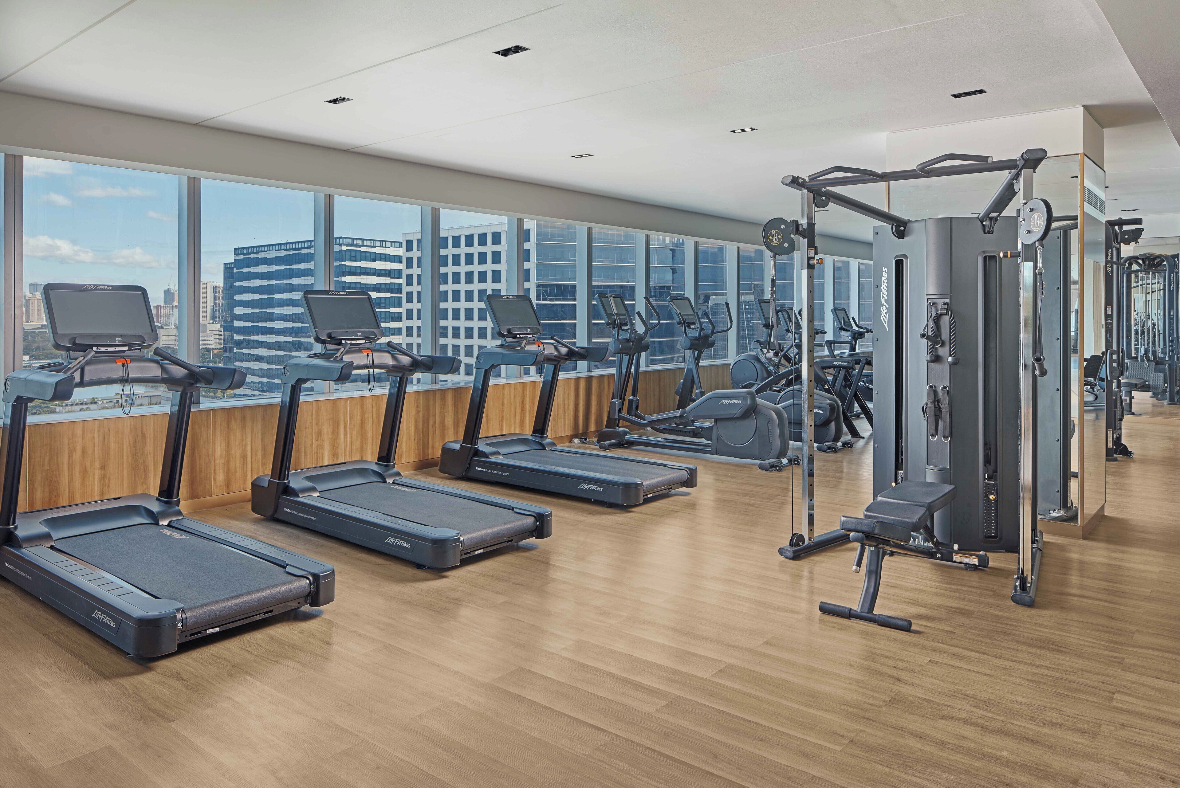 A modern gym with treadmills, elliptical machines, and weight equipment. Large windows offer views of a cityscape.
