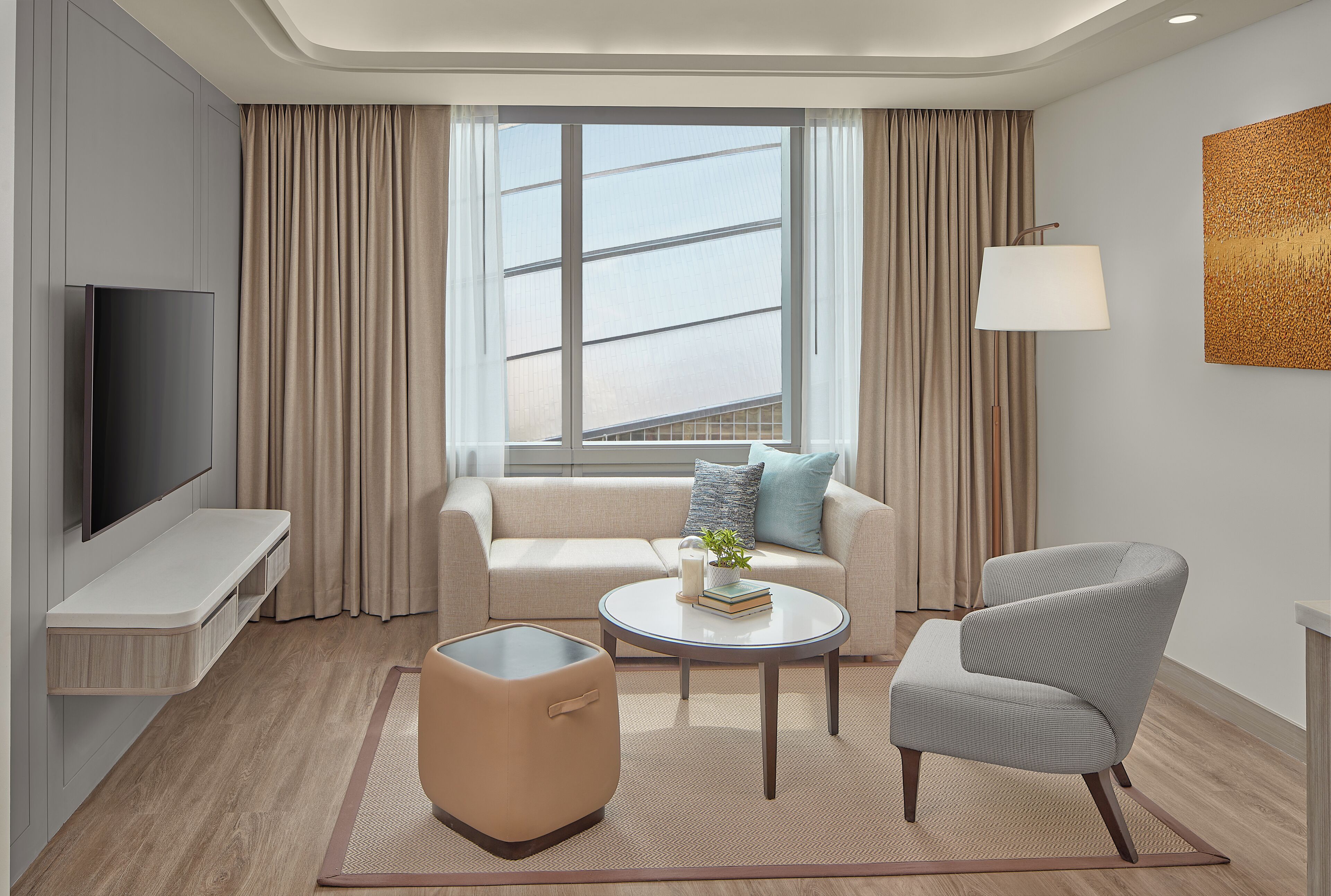 Una habitación de hotel moderna con sofá, sillón, mesa de centro, televisor y un amplio ventanal con cortinas.