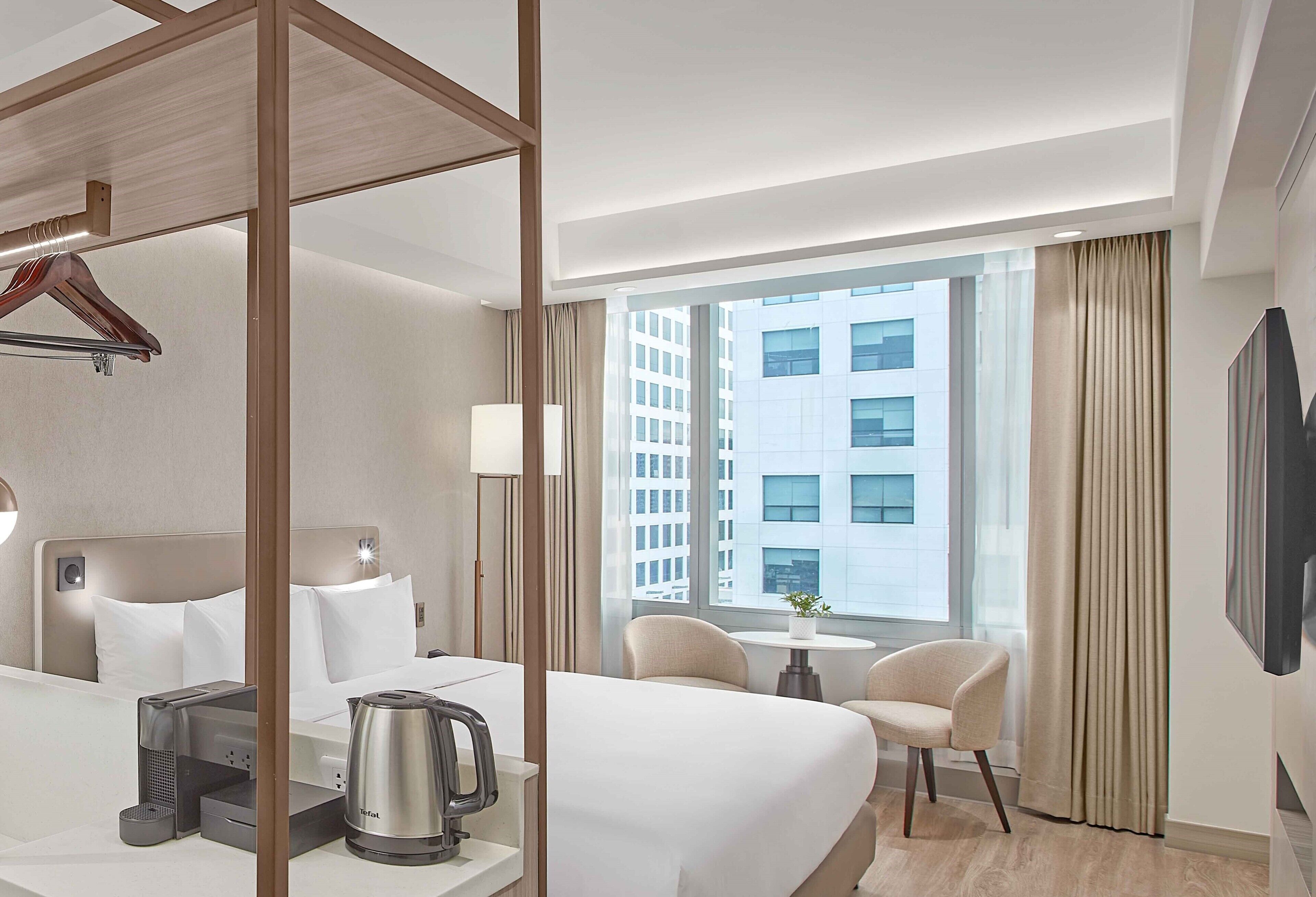 A hotel room with a bed, bedside table, kettle, and a view of a cityscape through large windows.