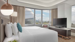 A modern hotel room with a large bed, a TV, a desk, and a view of a cityscape through large windows.