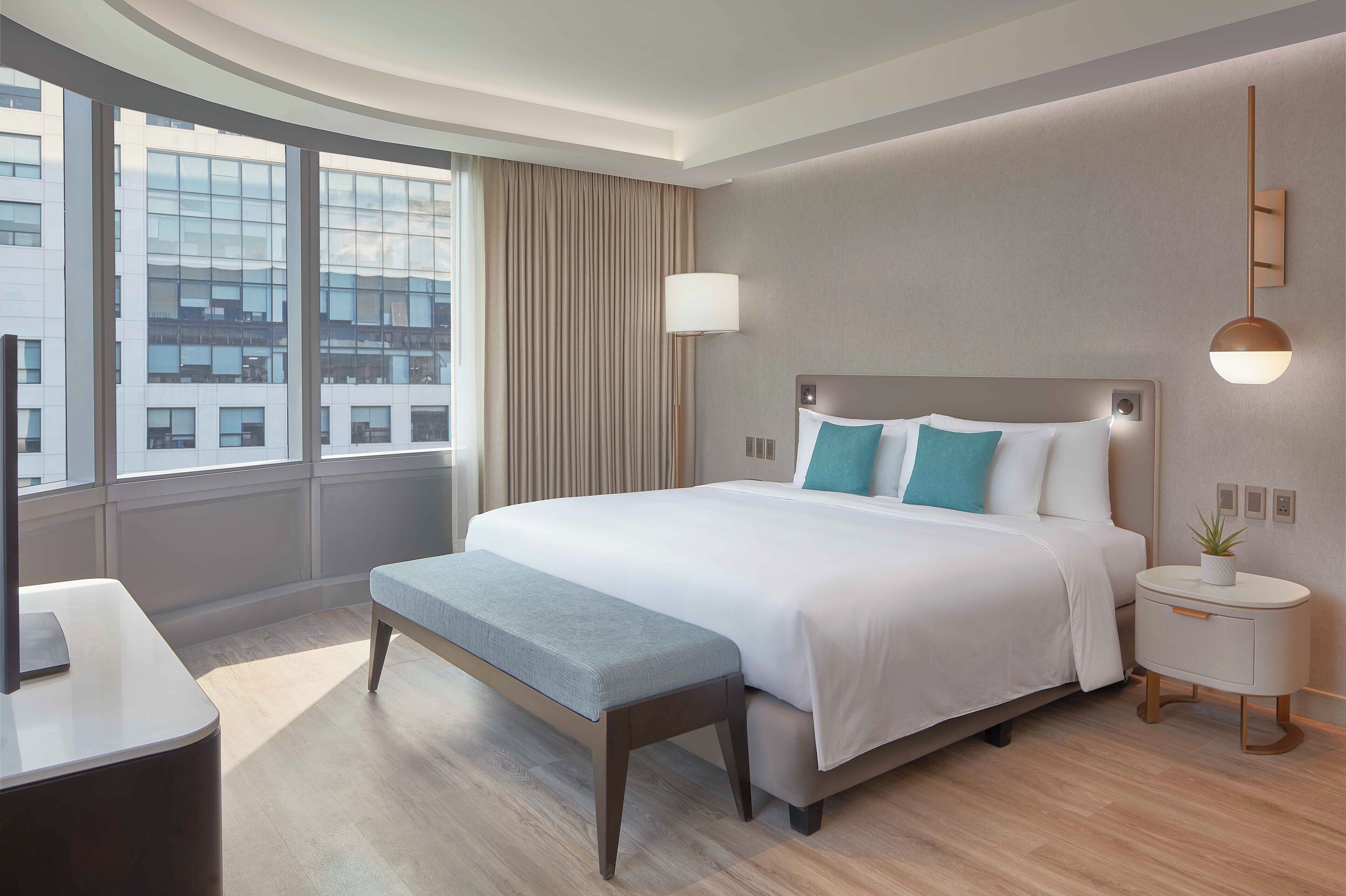 A modern hotel room with a large bed, a bedside table with a lamp, a bench, and a view of the city through large windows.