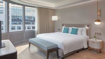 A modern hotel room with a large bed, a bedside table with a lamp, a bench, and a view of the city through large windows.