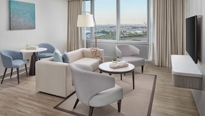 A modern hotel room with a large window, a flat-screen TV, and a seating area with two sofas and two armchairs.