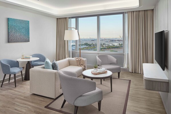 Una habitación de hotel moderna con un ventanal grande, un televisor de pantalla plana y una zona de estar con dos sofás y dos sillones.
