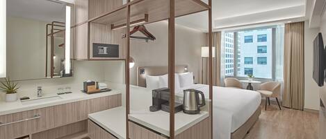 Een moderne hotelkamer met een groot bed, een nachtkastje met een waterkoker, een bureau met een telefoon en een plant, een grote spiegel en uitzicht op de stad door het raam.