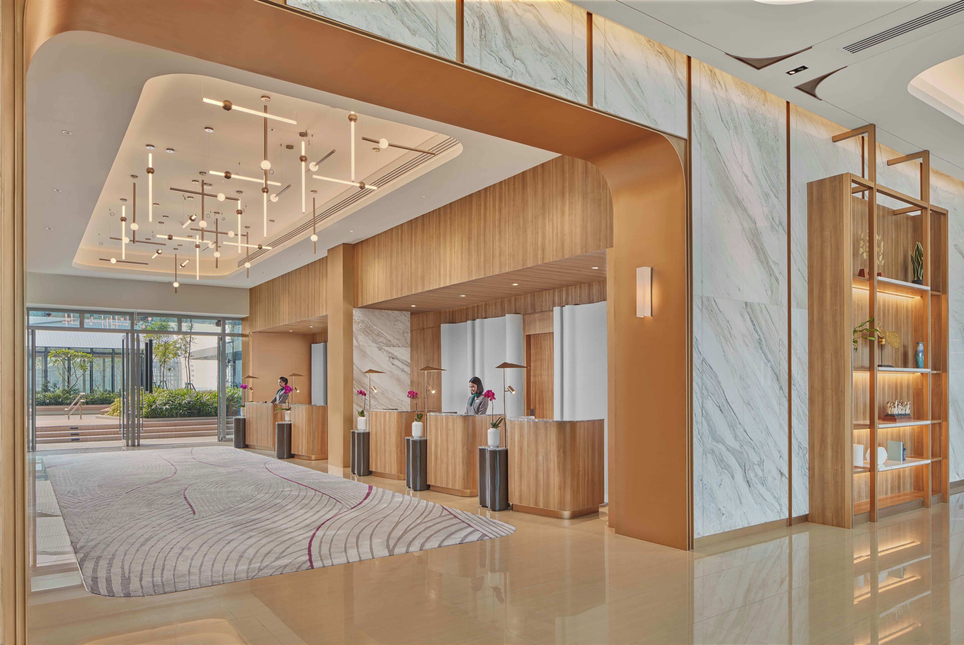 A modern hotel lobby with a reception desk, wooden paneling, marble walls, and a patterned rug.