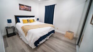 Een slaapkamer met een bed, nachtkastje, lamp, blauw gordijn en een klein mandje.