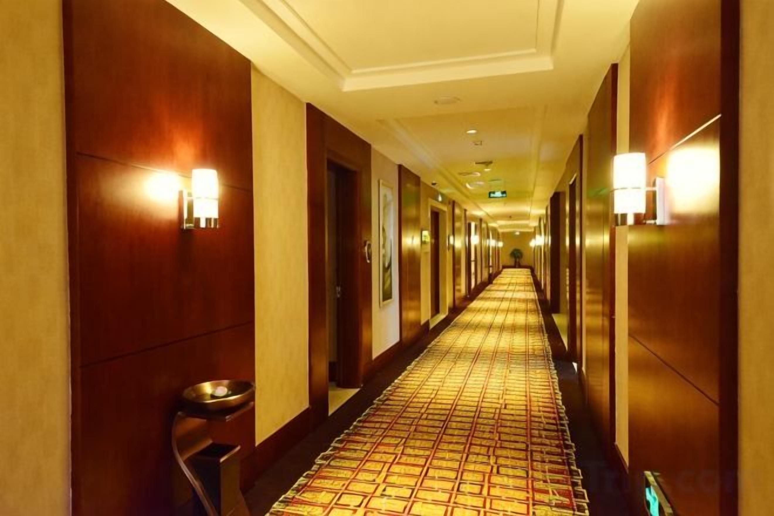 Corredor de hotel com carpete estampado, paredes de madeira e luminárias fixadas na parede.