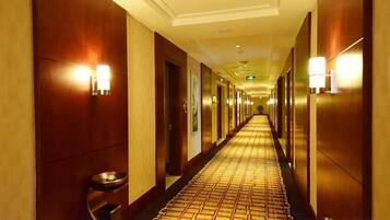 Corredor de hotel com carpete estampado, paredes de madeira e luminárias fixadas na parede.