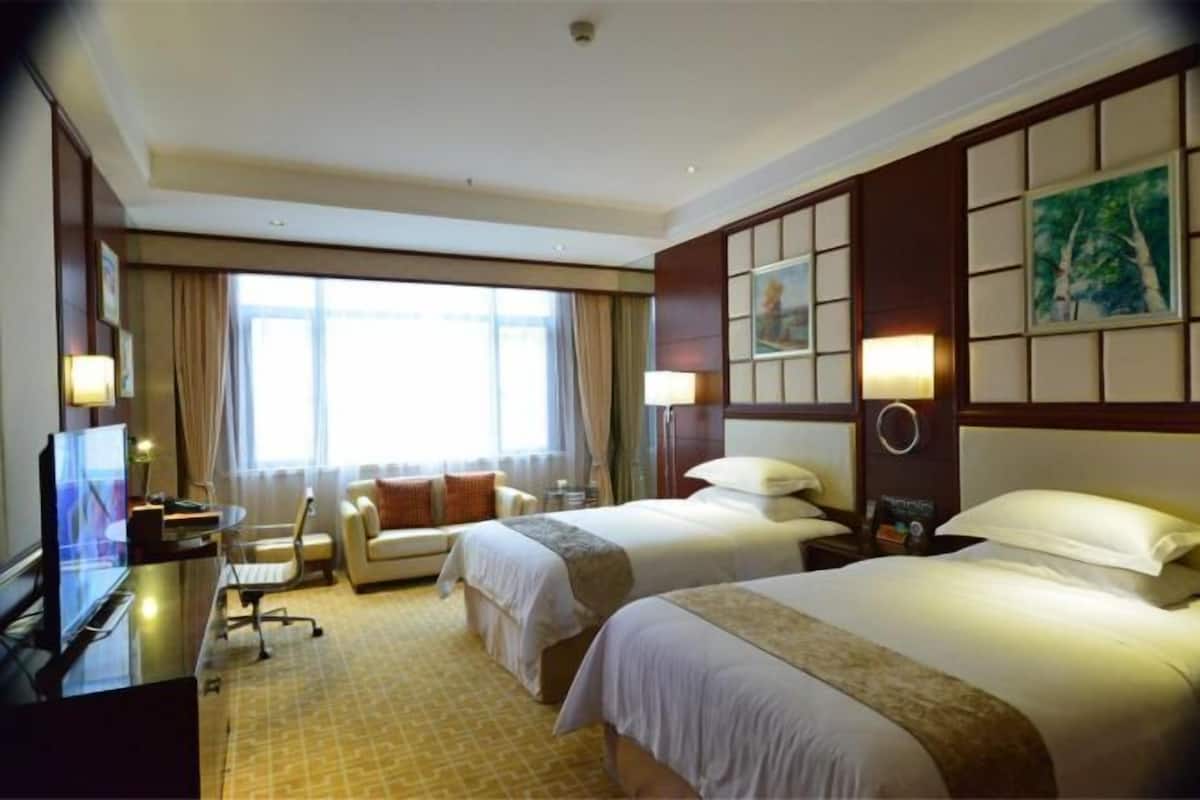 A hotel room with two beds, a desk with a TV, a sofa, and a large window.