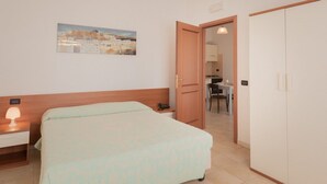 Ein Hotelzimmer mit Bett, Nachttisch mit Lampe, einem Bild an der Wand und Blick in den Küchenbereich.