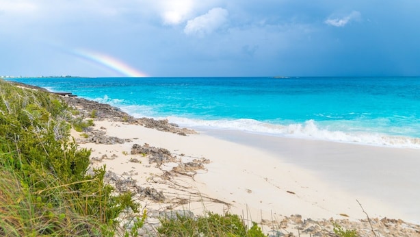 Een strand met helder turquoise water, een zichtbare regenboog en rotsachtig terrein.