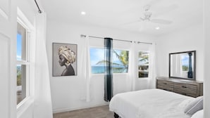 Een slaapkamer met een groot bed, een plafondventilator, een raam met uitzicht op zee en een schilderij aan de muur.