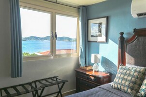 A bedroom with a large window offering a view of the sea and hills, a bed with patterned pillows, a bedside table with a lamp, and a small chair.