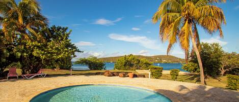 A swimming pool with a clear blue center, surrounded by a stone patio, palm trees, and a view of the sea and hills in the background.