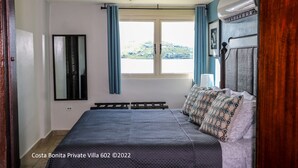 Een slaapkamer met een bed, een raam met gordijnen, een plafondlamp en uitzicht op een heuvel.