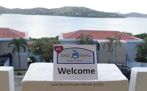 Welkomstbord voor Costa Bonita Private Villa 602, met uitzicht op zee en gebouwen op de achtergrond.