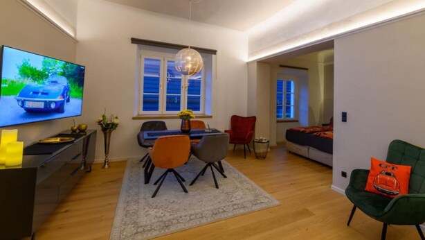 Una sala moderna con televisor de pantalla plana, una zona de comedor con sillas, un sofá y una cama en la habitación contigua.