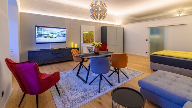 Un salon moderne doté d’un téléviseur à écran plat, d’une table à manger avec des chaises, d’un canapé et d’un lustre décoratif.
