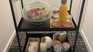 Ein Frühstückswagen mit Brot, Gebäck, einer Obstschale, einer Glasschale mit Schinken und Käse, einer Saftflasche, einer Kaffeetasse und verschiedenen Gewürzen.