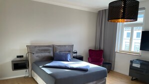 Ein modernes Schlafzimmer mit einem großen Bett, einem roten Sessel, einem Fernseher und einem Fenster mit Vorhängen.
