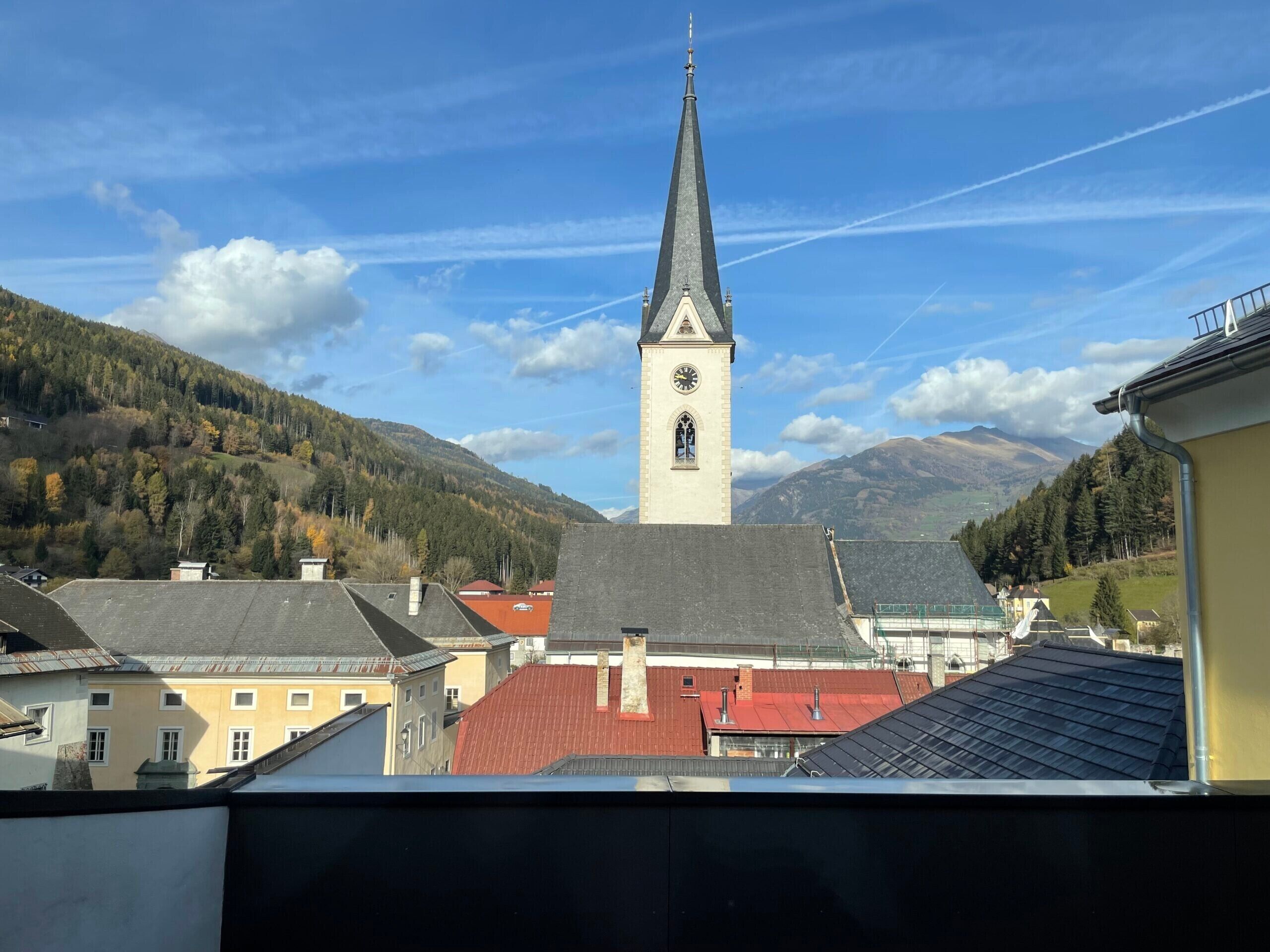 A church with a tall steeple and a clock, surrounded by buildings and mountains.